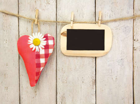 Wooden wall decorated with red fabric heart, marguerite and empty black boardの写真素材