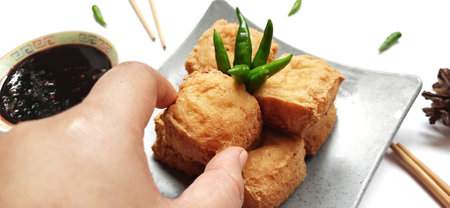 Fried milk tofu or Tahu susu goreng served on a plate and chopsticks with black sauce, green chili pepper on white background. Traditional snack from Indonesiaの写真素材
