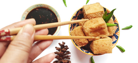 Fried milk tofu or Tahu susu goreng served on a plate and chopsticks with black sauce, green chili pepper on white background. Traditional snack from Indonesiaの写真素材
