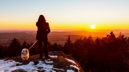 Young Man with his Dog on a walk against the setting winter sun in the mountains of Bavaria; Winter Sunset: Early March Winter Landscape Picture in Bavaria, Germanyの写真素材