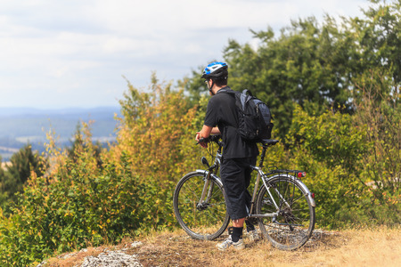 Biking in the Franconian Hills in Northern Bavaria. Caucasian Young man in black sports outfit with his Trekking bike on a rock, looking into the distance. Shot in High Summer August on an afternoonの写真素材
