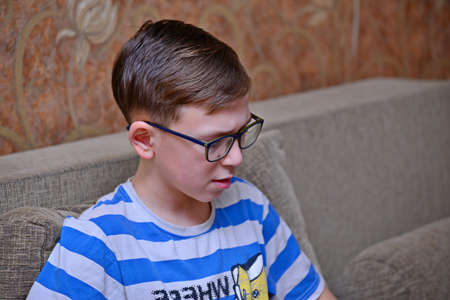 A boy in glasses with striped clothes is sitting on the couch and looking