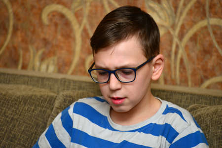 A boy in glasses with striped clothes is sitting on the couch and looking