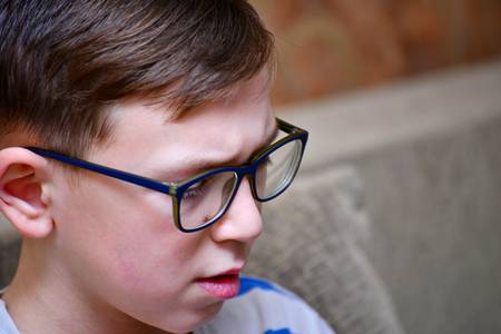 A boy in glasses with striped clothes is sitting on the couch and looking