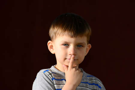 The boy holds a finger at the mouth. Portrait of a boy on a dark background in the studio, one side is lit up by more than anotherの写真素材