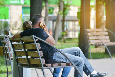 A man is sitting on a bench in a park talking on a smartphone on the streetの写真素材