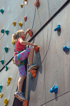The girl is blonde, climbs to the top on an artificial rock with the help of a safety rope, is engaged in rock-climbingの写真素材