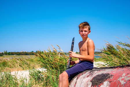 A boy is playing on a black clarinet sitting on an old wooden boat on the beachの写真素材
