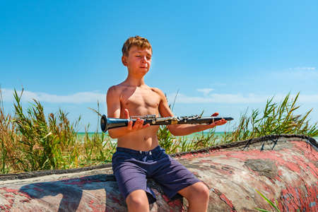 The boy holds a black clarinet in his hands, comprehending Zen sitting on an old wooden boat on the beachの写真素材