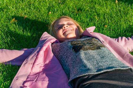 The girl in the pink coat is lying on the green grass in the park in autumn