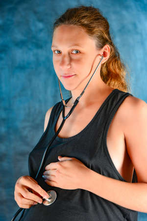 Sexy pregnant girl close-up, looks in to the lens in a black T-shirt and blue shorts, blue background, listens to his stomach through a stethoscopeの写真素材
