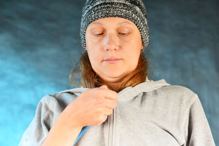 A girl in a knitted hat with red hair fastens a sports jacket on a blue background in the studioの写真素材