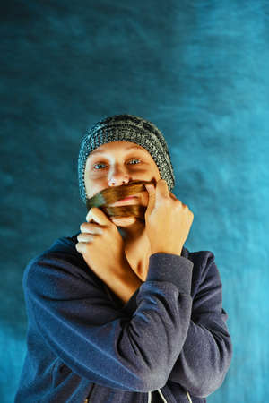 A girl in a knitted hat with red hair makes a mustache out of hair, on a blue background in the studioの写真素材