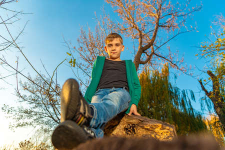 A boy in jeans is sitting on a log, a child is sitting on a big dry tree in a park, wide-angle close-up photo in the forestの写真素材