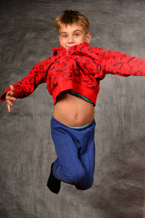 A boy in a red sweater and blue pants jumps and holds his hand up in the studio on a gray background