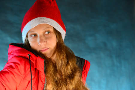 Lovely young girl in a red jacket and Santa Claus hat with red hair on a blue background in the...