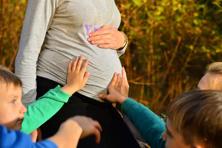 Young children sit next to a pregnant woman and hold their hands on their mother's belly in the...