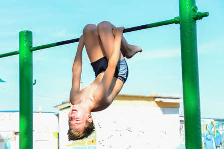 A boy in shorts goes in for sports on a horizontal bar on the street. Does various exercises,...