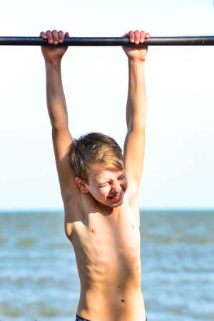 A boy in shorts goes in for sports on a horizontal bar on the street. Does various exercises,...
