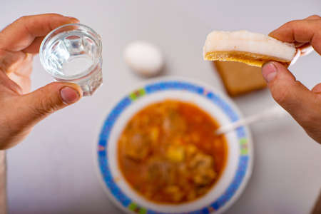 A man holds a pile of alcohol in his hands and eats black bread with bacon and red soup while...