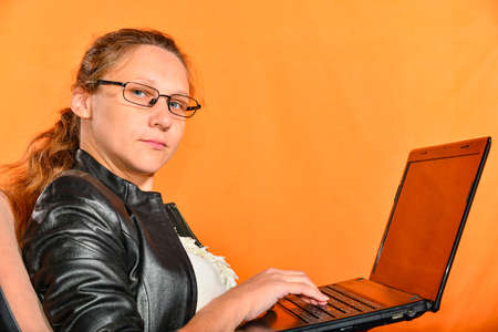A young girl in a black jacket and glasses holds a laptop and looks into the camera.