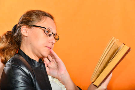 Charming surprised girl in glasses sits and holds a brown book on an orange background.