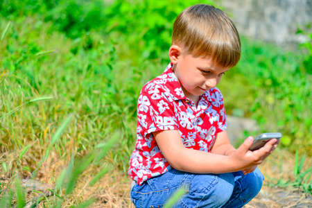 A boy with a smartphone sits on the grass and looks at the screen of the phone.