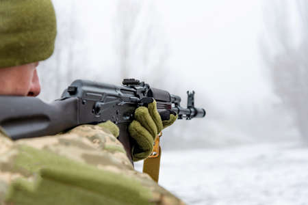 A military soldier takes aim with an automatic rifle and holds it in his mittens in winter.