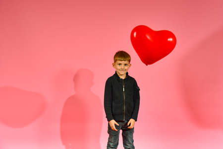 A boy throws and catches a heart-shaped balloon on a pink background in the studio.
