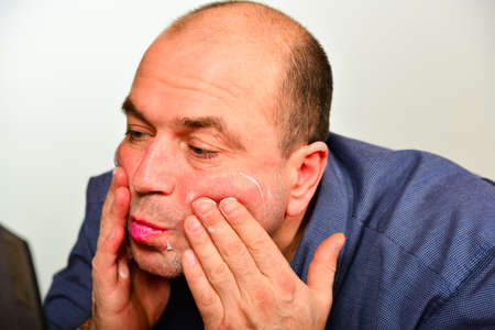 An adult and unshaven man in a blue shirt applies a mask of cream on his face, the concept of...