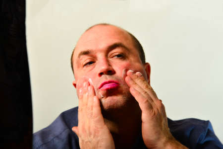 An adult and unshaven man in a blue shirt applies a mask of cream on his face, the concept of...