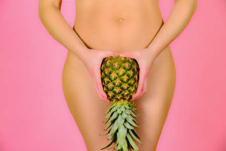 A young and sexy girl is holding a pineapple near the feet, on a pink background.の写真素材