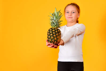 A lovely girl holds out her hand with a pineapple, on a yellow background in the studio.の写真素材