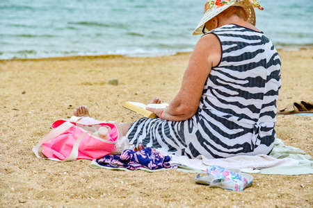Grandmother reads a book on the beach sitting on the sand by the sea.の写真素材