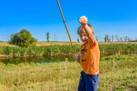 A fishing boy on a river holds a fishing rod, a child caught a fish and pulls it out of the water on a hook.の写真素材