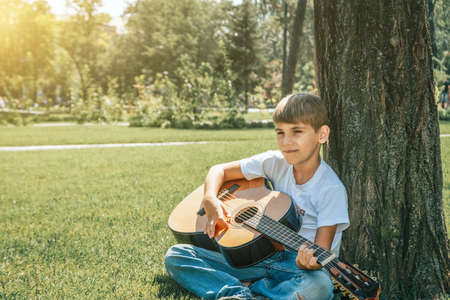 A boy with a guitar sits under a tree, sings songs and enjoys nature.の写真素材