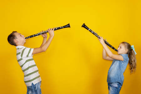 A boy and a girl play the clarinet and stand against each other, on a yellow background.