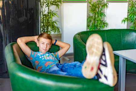 The boy sits on a green armchair, legs topped and enjoys the rest, looking at the camera.の写真素材