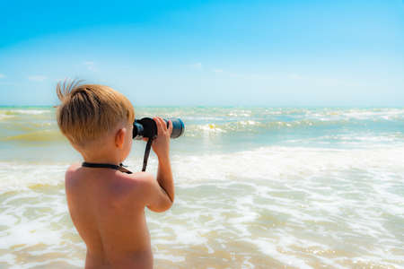 A boy with binoculars looks at the sea against a blue sky.