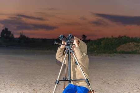 A boy looks through binoculars on a tripod against the background of an evening sunset.の写真素材