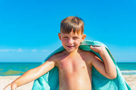 The boy on the beach hides with a towel and wipes himself from sea water after swimming in the sea.の写真素材