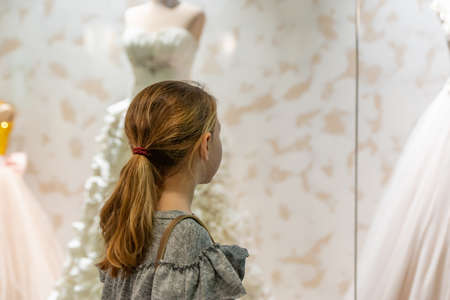A young girl, a teenager, stands near the window of a wedding salon and looks at wedding dresses, introducing family life.の写真素材