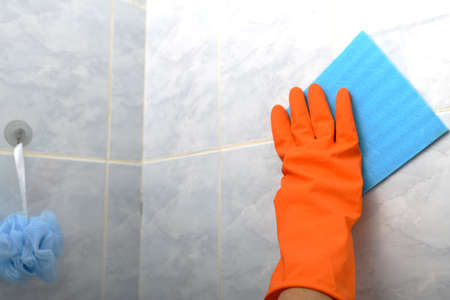 A woman wipes a tile with a blue napkin in a bathroom in orange protective glovesの写真素材