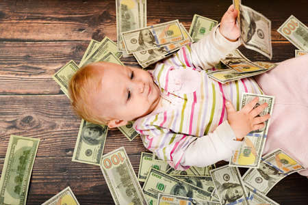 A small child, a girl lies on a wooden background, dollars are falling from above on a child. The concept of wealth and financial independenceの写真素材