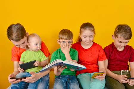 Children sit next to each other and look at the phone, while one boy in glasses sits with a book. The concept of online learning at homeの写真素材