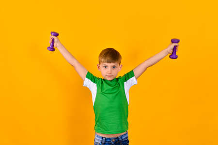 A boy in a green sports shirt holds his hands with dumbbells on the sides, a sports child on a yellow backgroundの写真素材