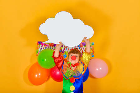 A baby clown on a birthday background, holding a white sign in the form of a cloud for inscription...