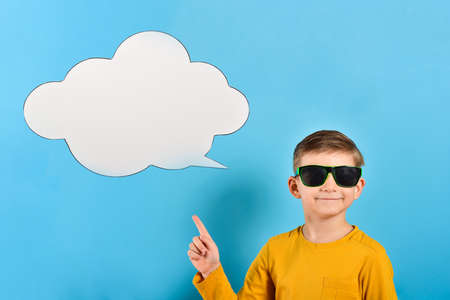 Joyful and smiling boy in glasses shows a finger at a white cloud for text or advertising...