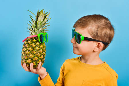 The boy with glasses holds a pineapple in his hand with glasses, and they look at each other. Baby with pineapple on a blue background. Children, sun, summer and food.の写真素材