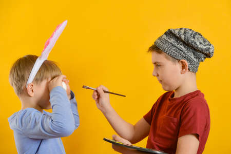 A young artist in a hat with a palette in his hand paints Easter eggs, the children are preparing for the holiday of Easter.の写真素材